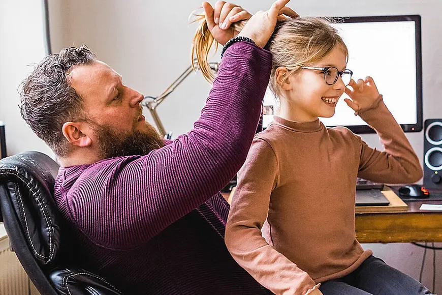 Ein Vater macht seiner Tochter einen Zopf, im Hintergrund ein Computer auf einem Schreibtisch 