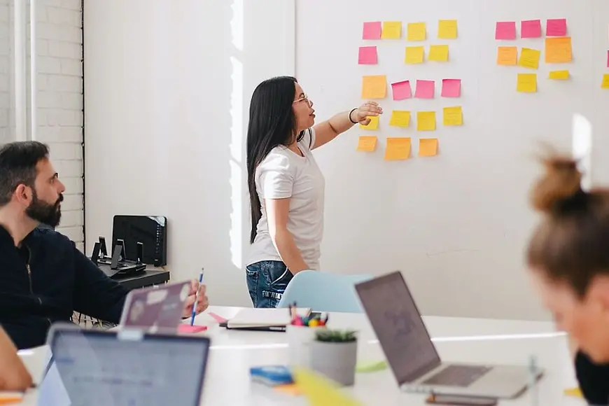 Eine Frau zeigt auf Klebezettel an der Wand, vier Personen sitzen am Tisch vor ihren Laptops