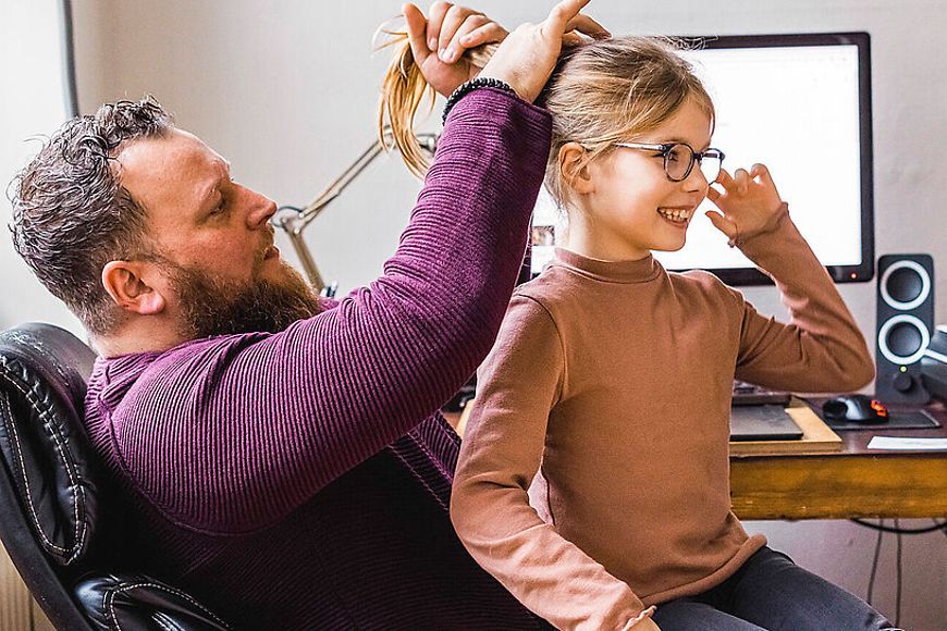 Ein Vater macht seiner Tochter einen Zopf, im Hintergrund ein Computer auf einem Schreibtisch 