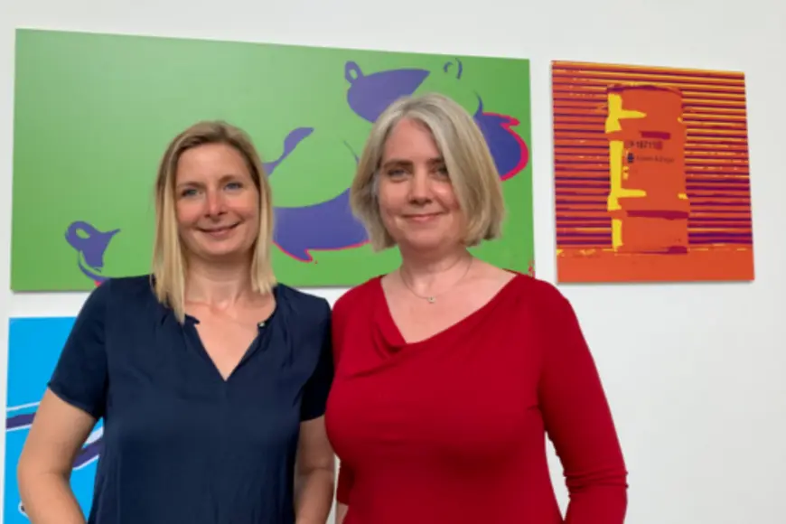 zwei Frauen schauen in die Kamera und stehen vor einer Wand mit Bildern