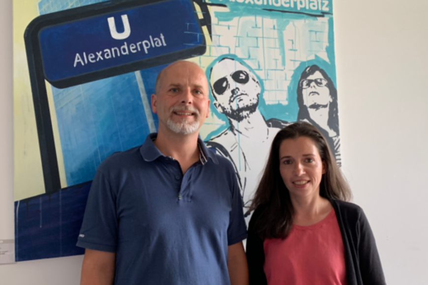 Ein Mann und eine Frau schauen in die Kamera nd stehen vor einem Bild mit der Aufschrift "U Alexanderplat"