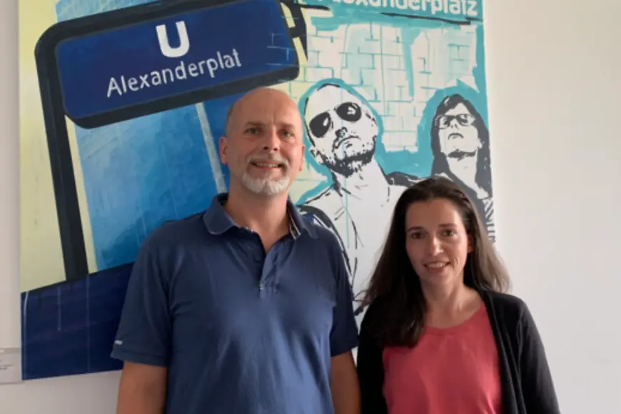 Ein Mann und eine Frau schauen in die Kamera nd stehen vor einem Bild mit der Aufschrift "U Alexanderplat"