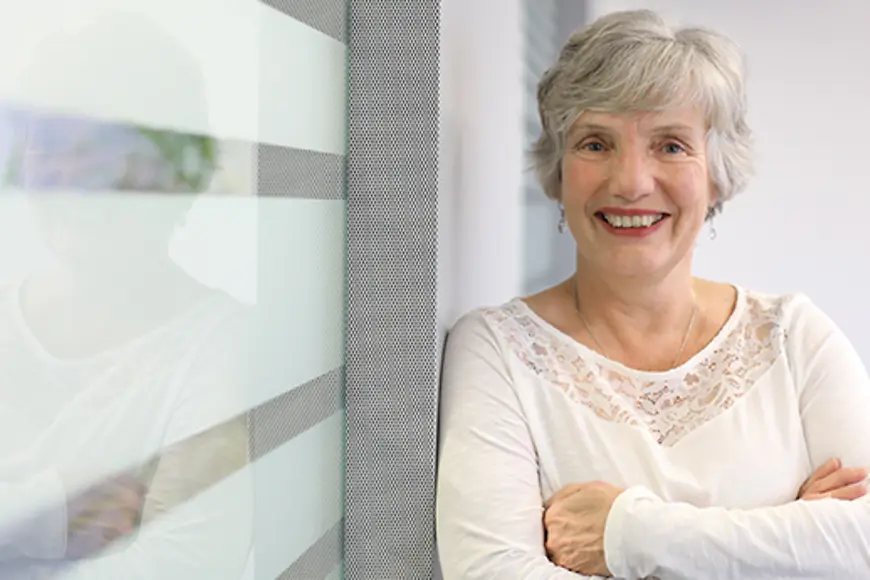 Eine ältere Frau lehnt an einer Glaswand und schaut in die Kamera