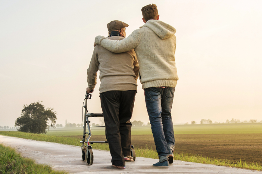 Ein älterer Mann mit Rollator, ein jüngerer Mann legt den Arm um ihn, beide laufen in der Natur